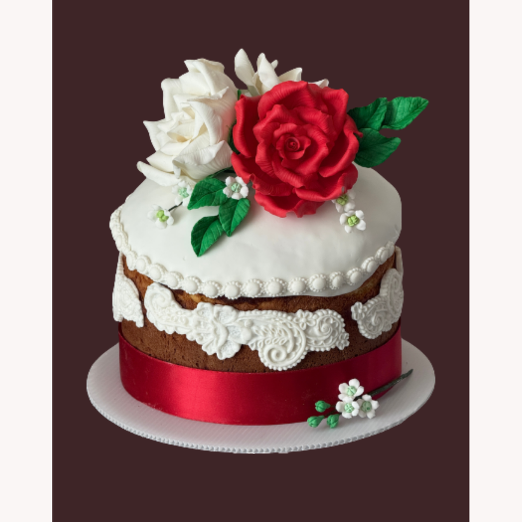 A decorated two-tier cake with white icing, a red satin ribbon, white lace trim, a large red rose, white flowers, and green leaves on a white board against a dark brown background.